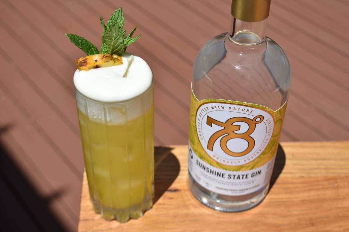 Frothy cocktail garnished with mint and pineapple beside a bottle of 78 Degrees Sunshine State Gin