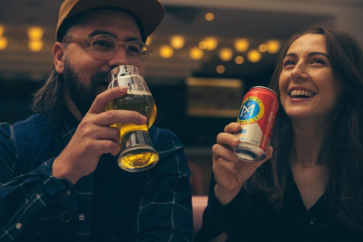 Couple at the bar, one sipping a pale ale, the other laughing with a Mismatch can in hand